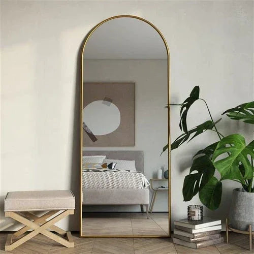 Full-length mirror with gold frame in a bedroom setting with a plant and books.